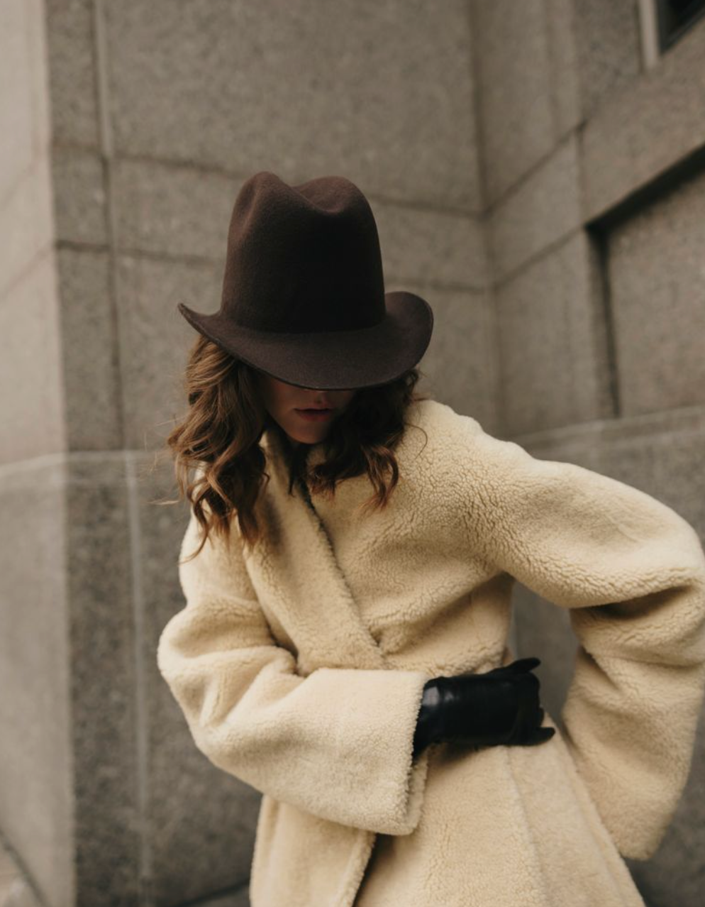Person wearing a beige coat and brown hat against a gray building.