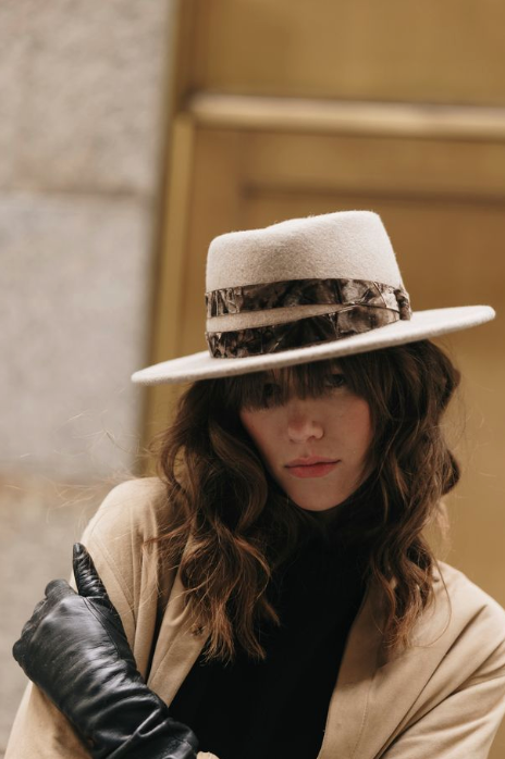 Person wearing a white hat with a band, beige coat, and black gloves against a neutral background
