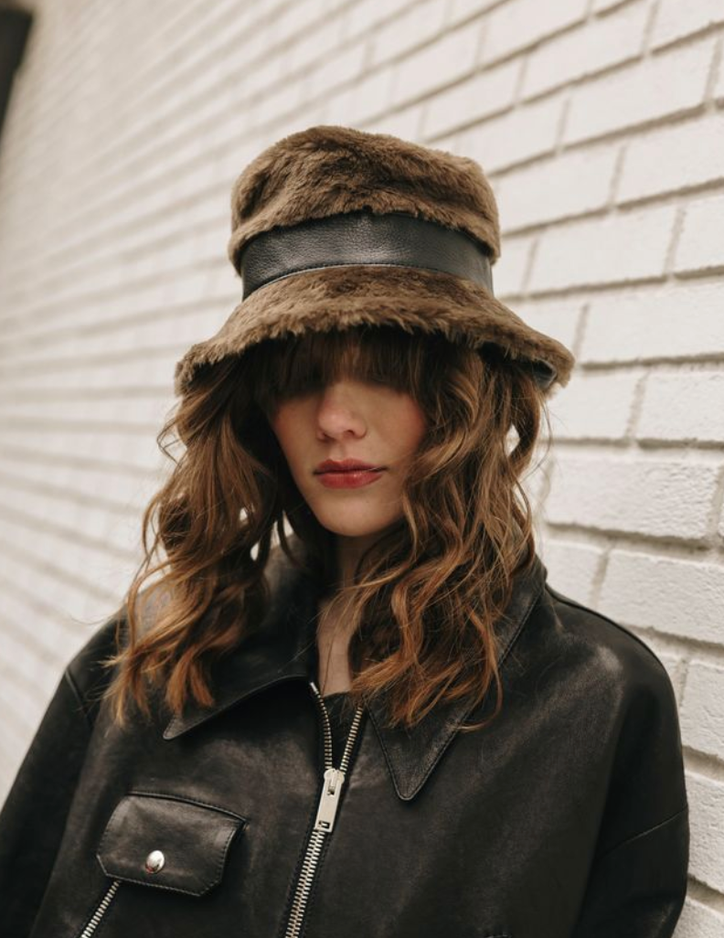 Person wearing a fur hat and black leather jacket against a light-colored wall.