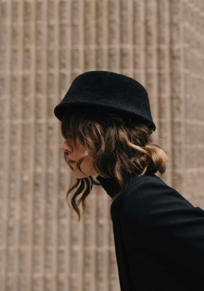Person wearing a black bucket hat and coat against a textured wall.